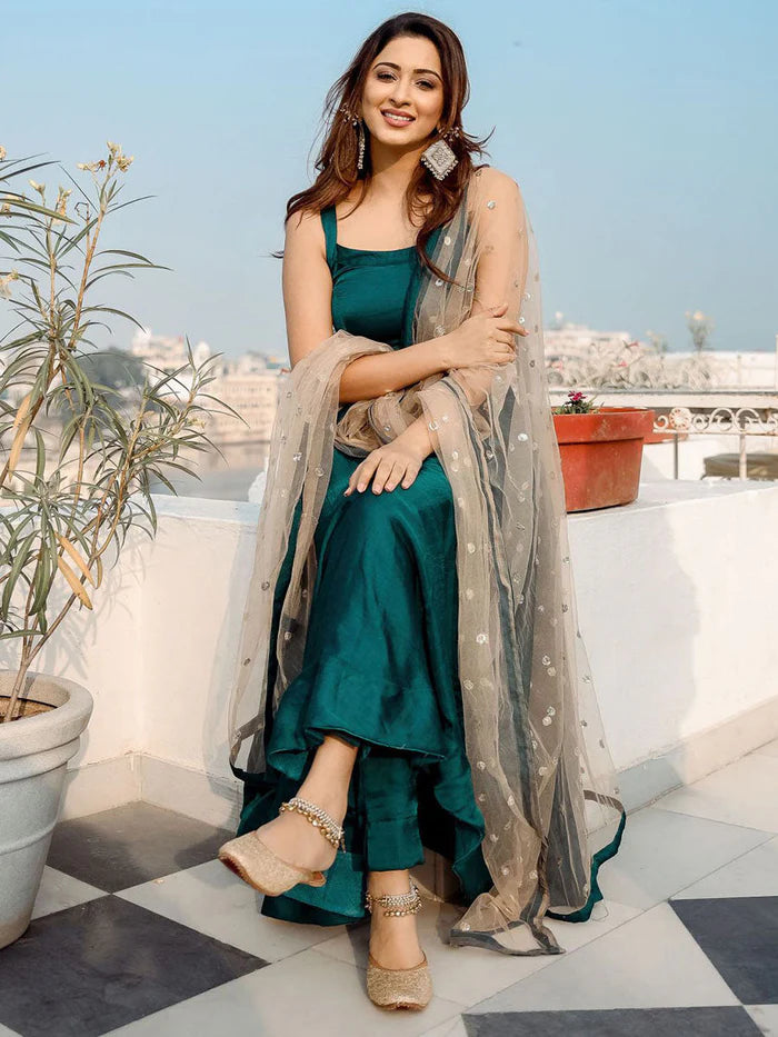 Woman in a teal dress with a beige dupatta on a rooftop with plants and a cityscape in the background